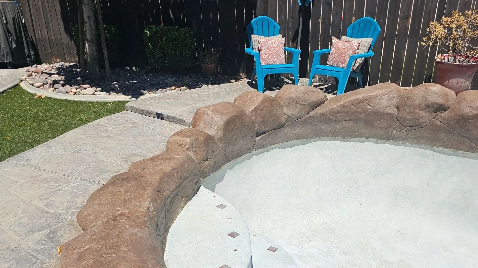 Backyard with a stone-edged pool, green lawn, and two blue chairs on a patio.