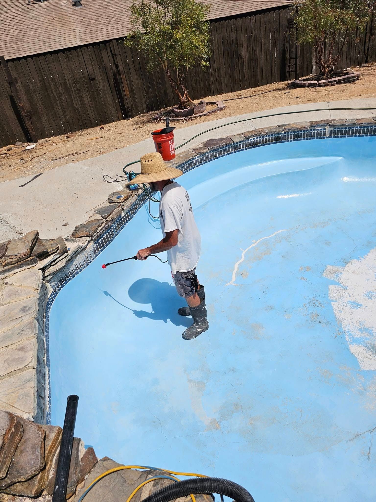 Person standing in an empty blue swimming pool using a hose or pressure washer to clean it