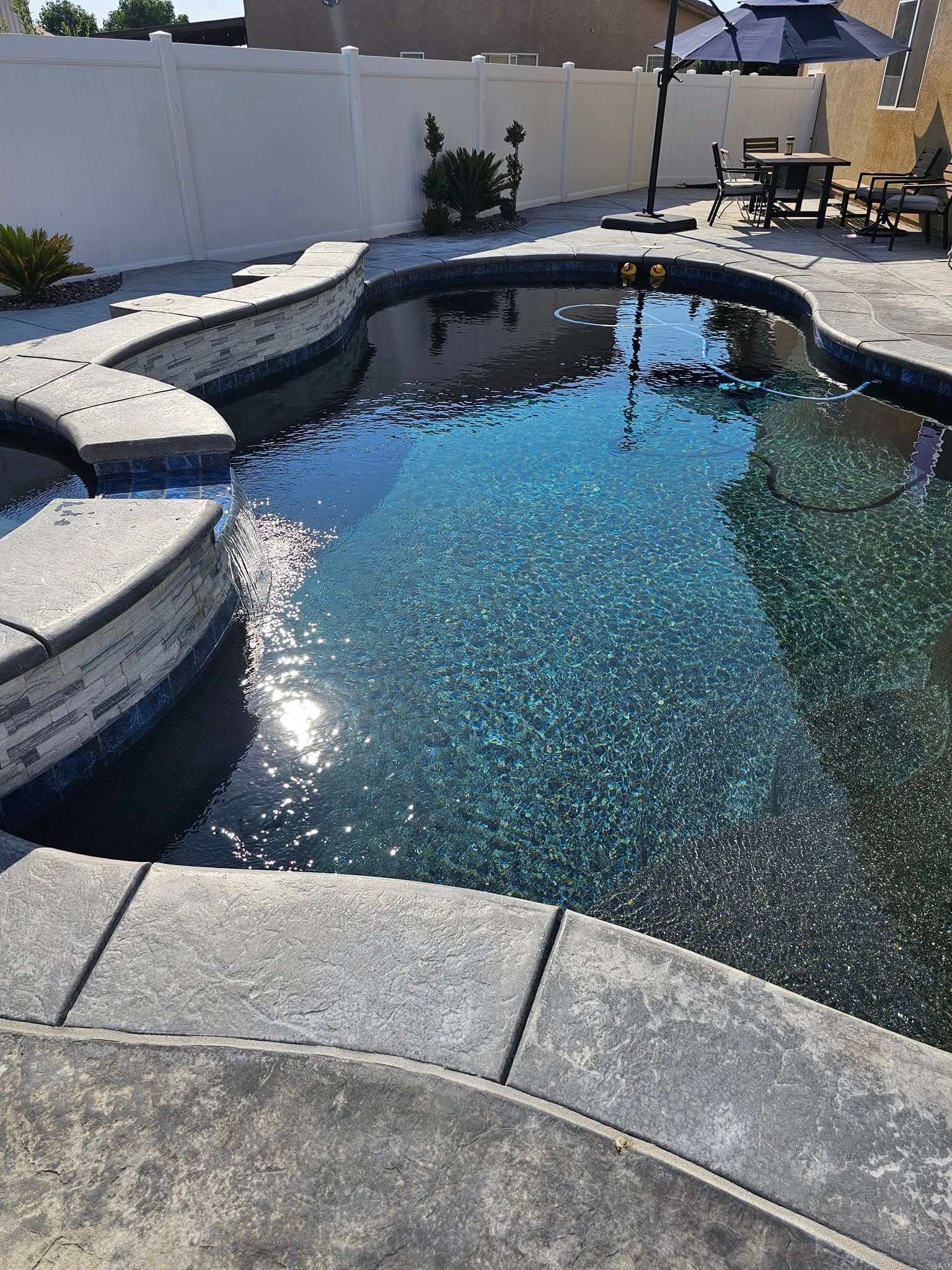 Backyard swimming pool with blue water, stone coping, and patio seating in the background