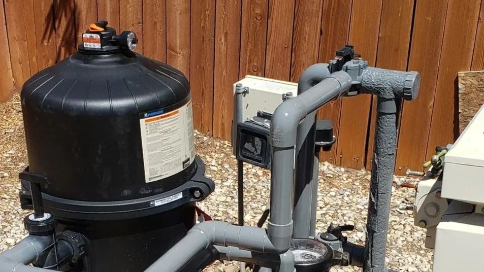 Black pool filter and gray pipes beside a wooden fence outdoors