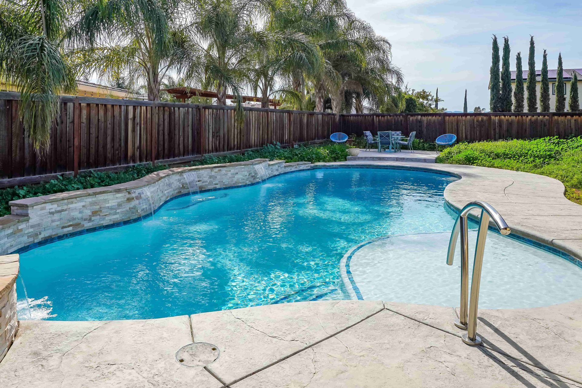 Backyard swimming pool with blue water, stone deck, and wooden fence under a clear sky