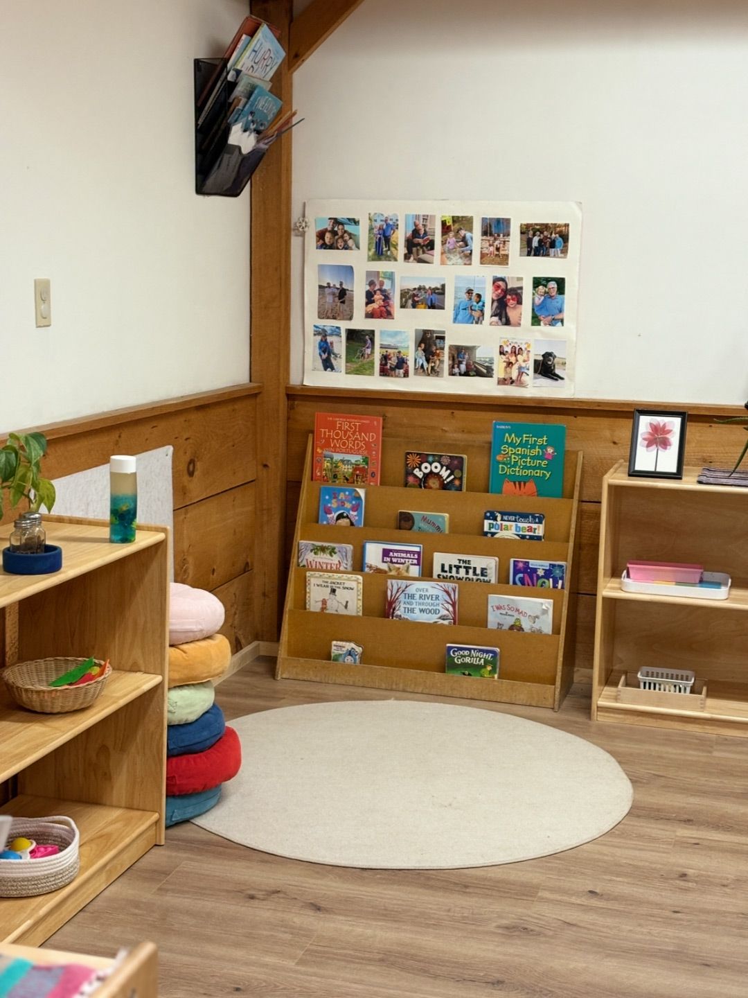A toddler Montessori reading area featuring a low book display, family photo board, and a circular floor rug.