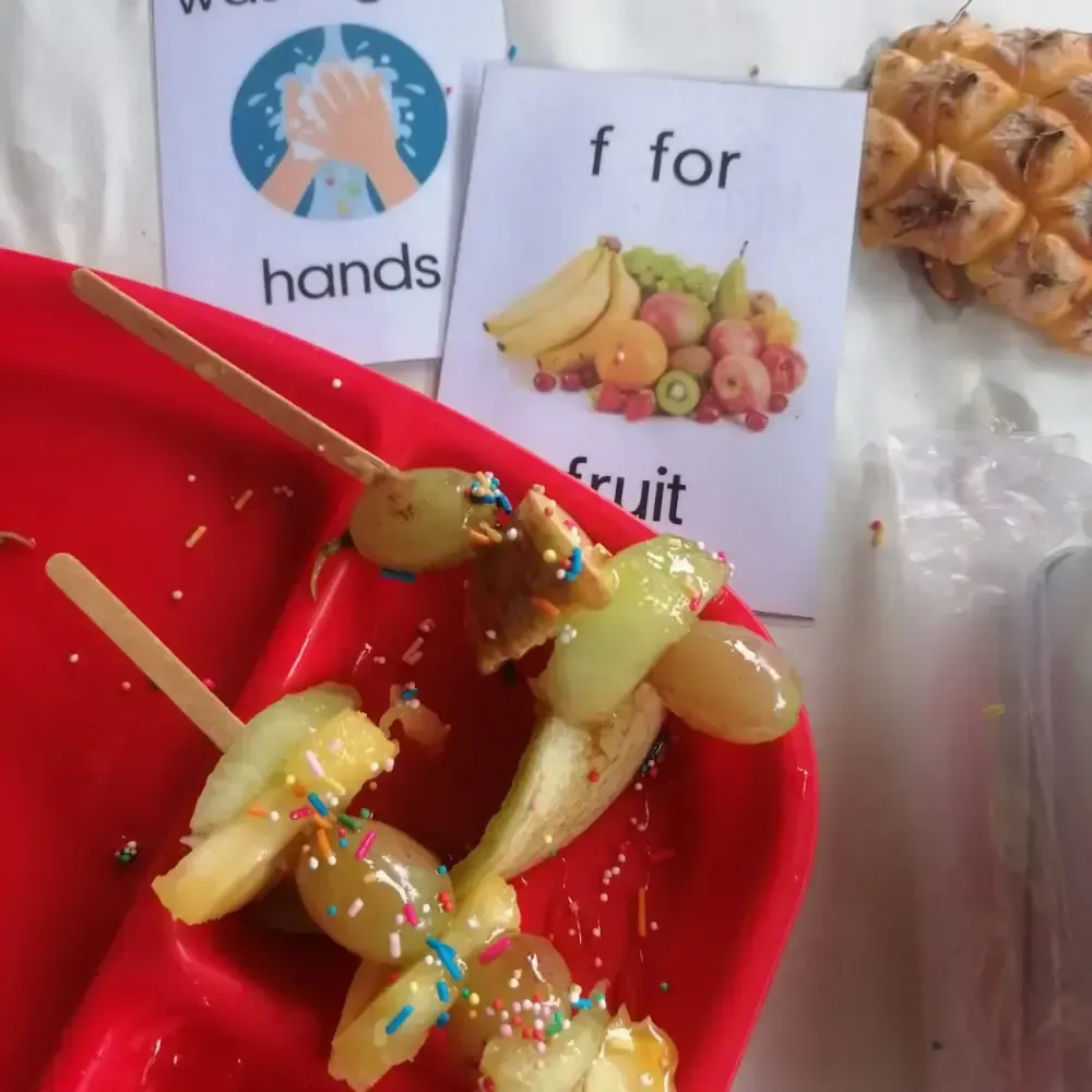 Children’s fruit snacks with apple slices, caramel, and sprinkles on red plates, beside alphabet cards.