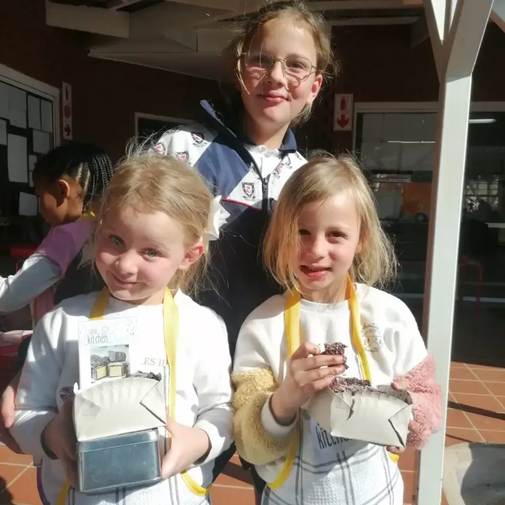 Three children outdoors, smiling and holding small boxes or craft projects in sunlight.