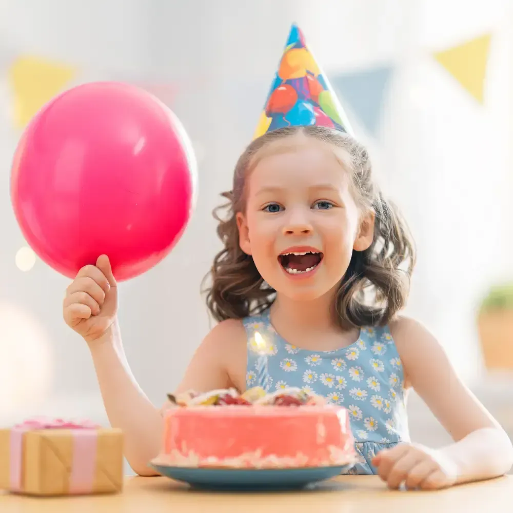 Child at a birthday table holding a pink balloon, smiling beside a cake and gift box