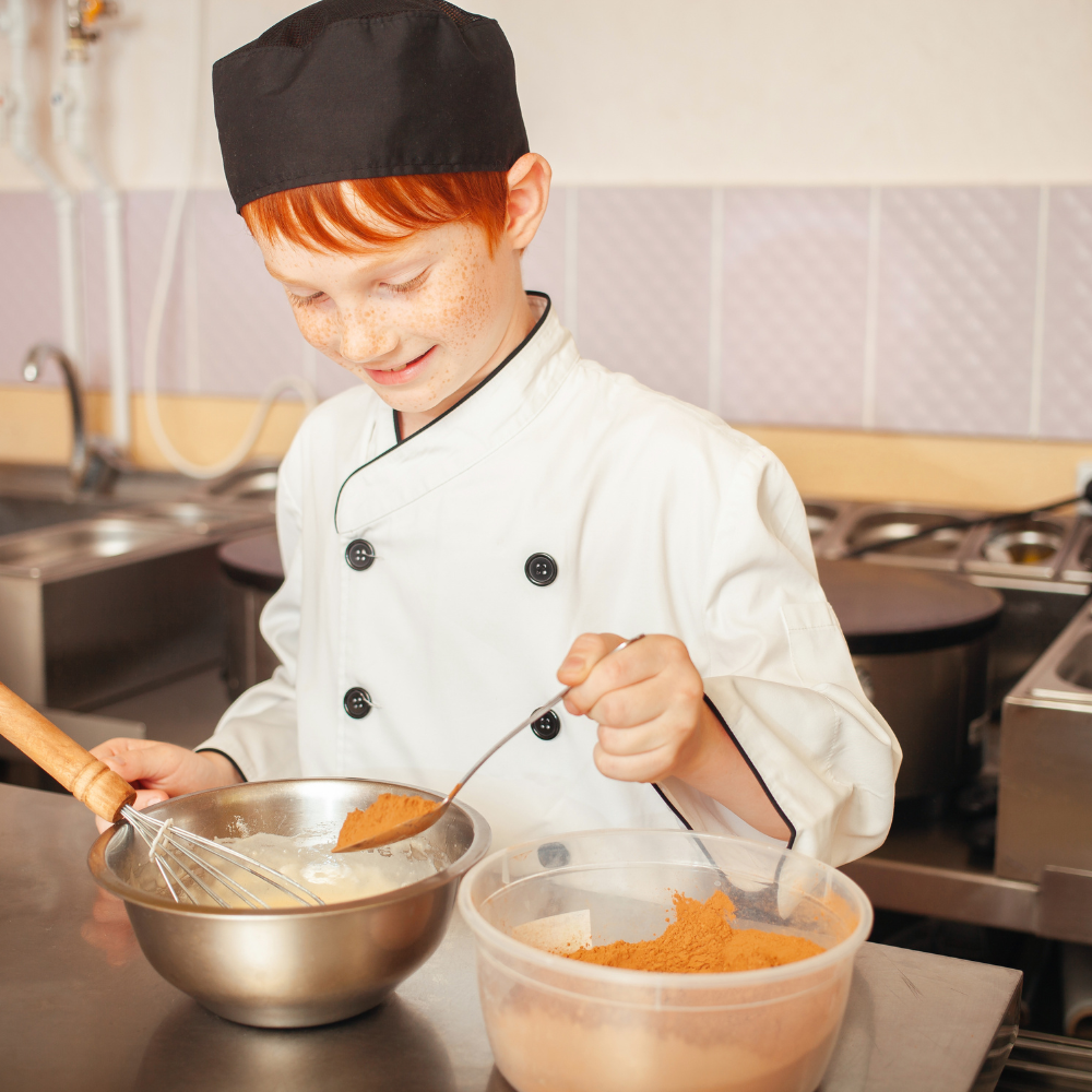 Young chef in a white coat and black hat whisking batter in a kitchen with a bowl of orange ingredients