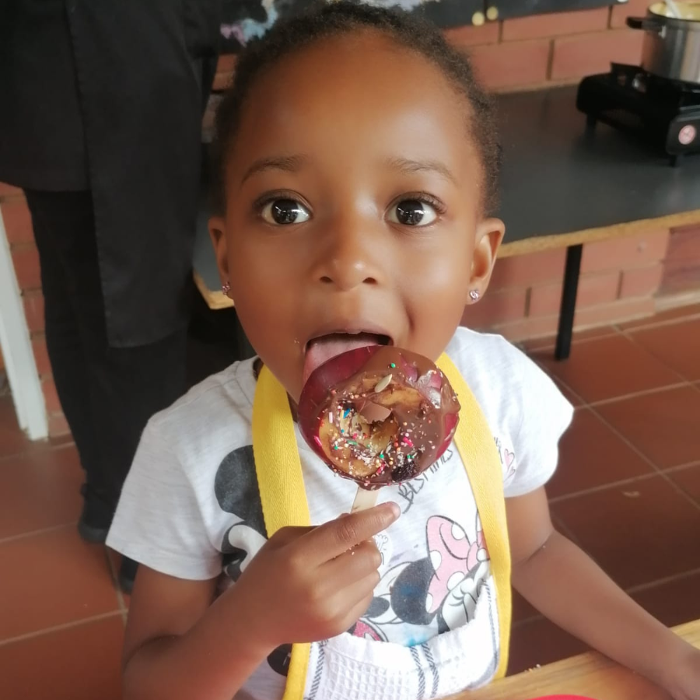 Child holding and licking a sprinkled lollipop indoors, wearing a white shirt and yellow suspenders.