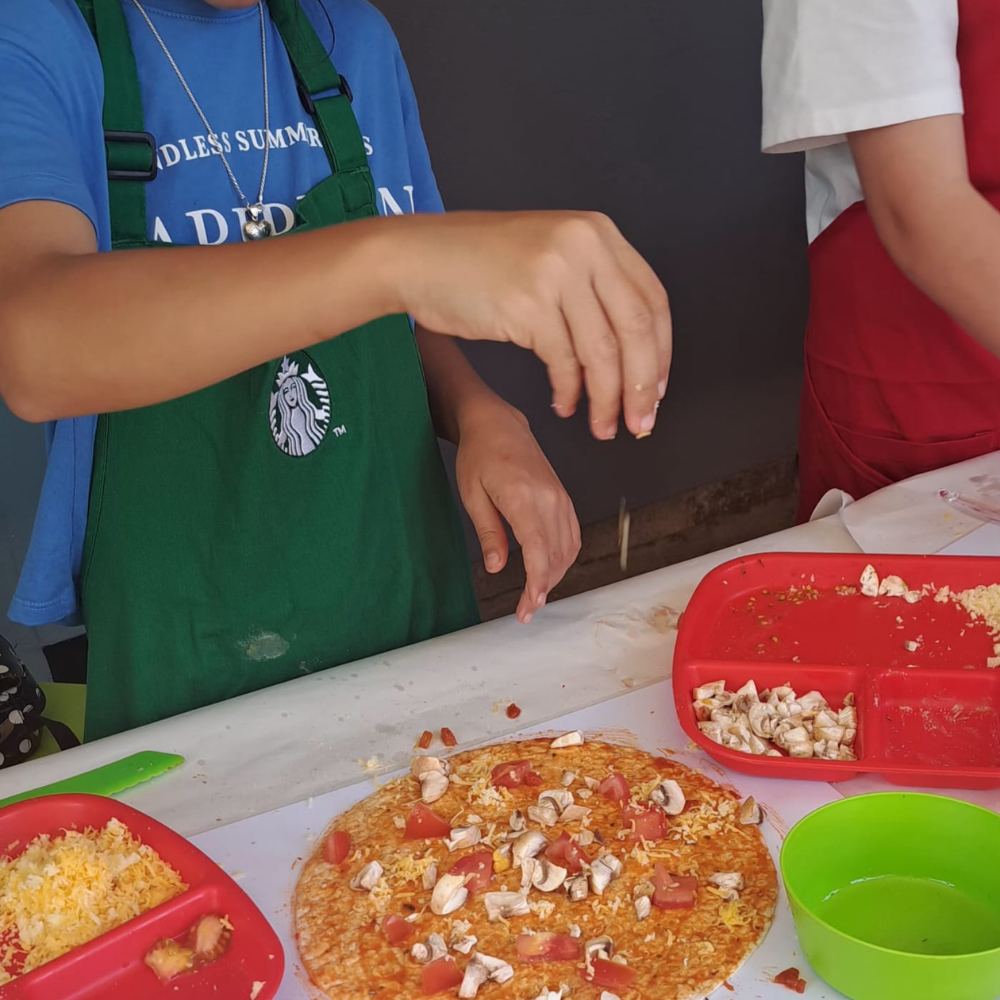 Children assembling a colorful pizza with toppings at a table, using red and green trays