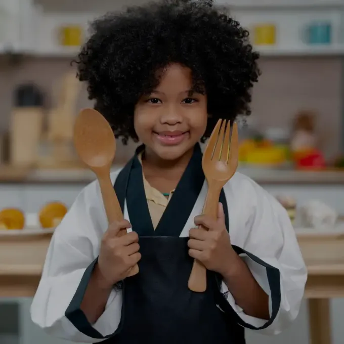 Child in a kitchen apron holding a wooden spoon and fork, smiling at the camera.