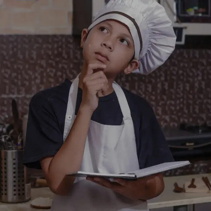 Child chef in a white hat and apron thinking while holding a notebook in a kitchen