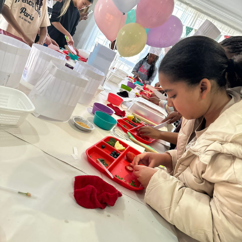 Child making a colorful craft at a table with balloons in the background