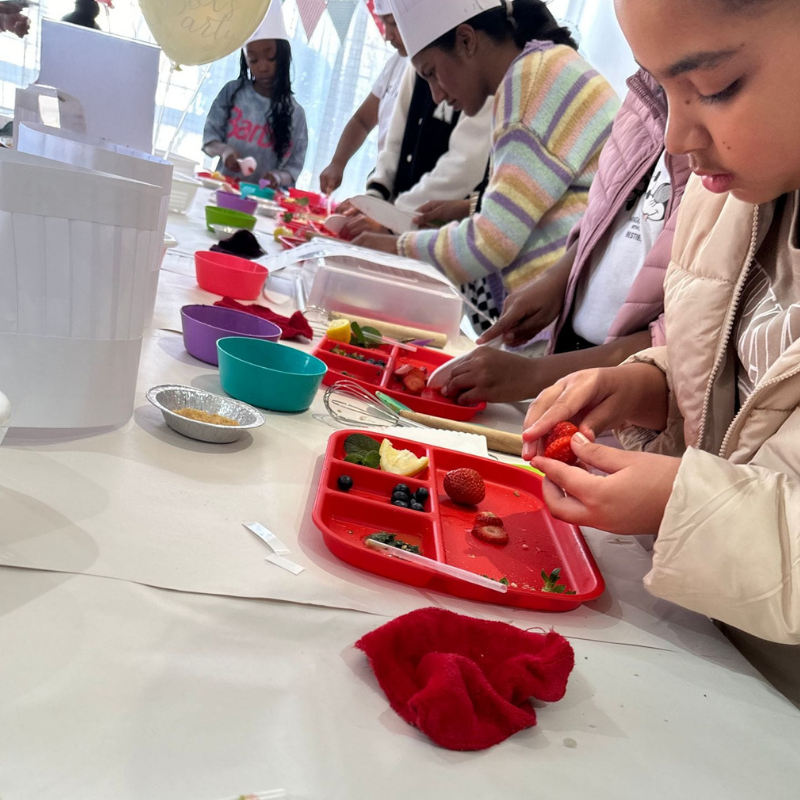 People making crafts at a table with red trays, small bowls, and colorful materials in a bright room