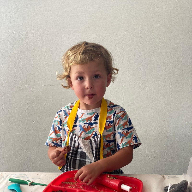 Child at a table holding a yellow ribbon and a pencil, with a red toy and papers in front of them.