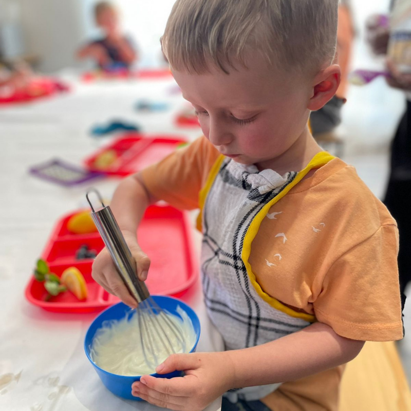Child whisking batter in a blue bowl at a table with red trays and toys.