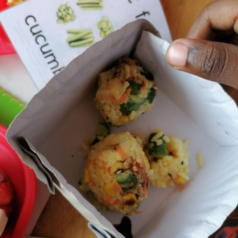 Hand holding a paper tray with three small savory snack balls inside, with a red item in the background