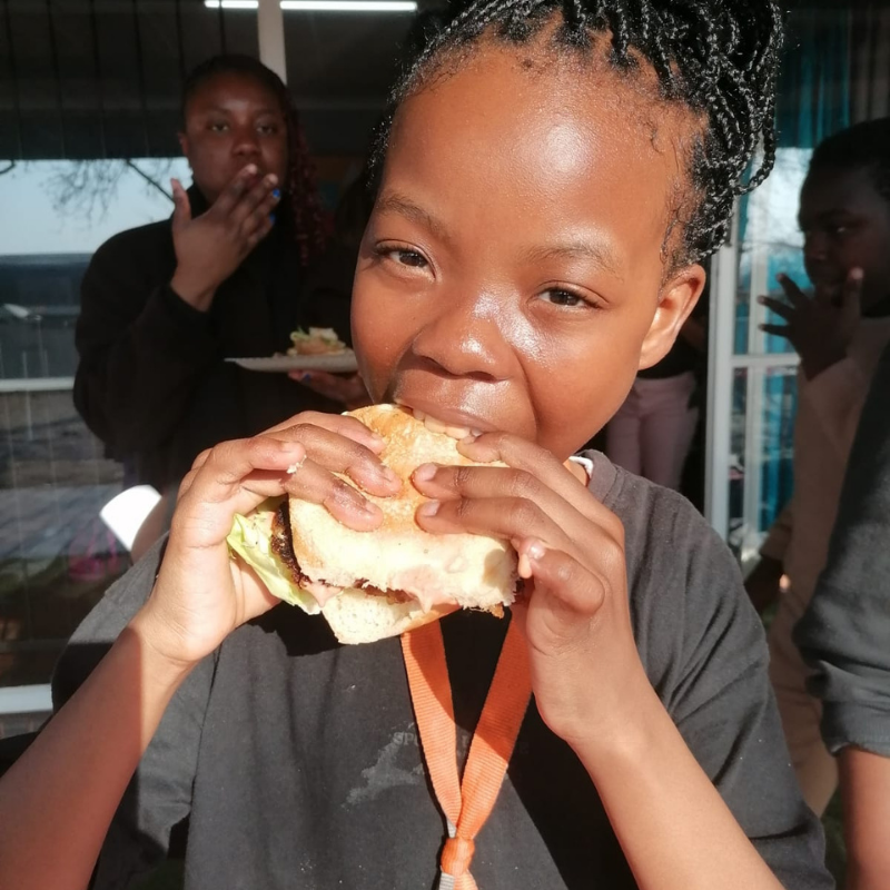 Child eating a sandwich outdoors in bright sunlight, with two people blurred in the background