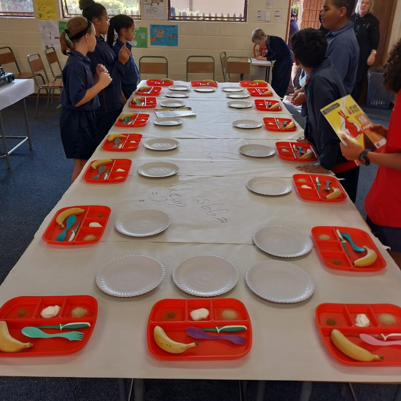 Children standing around a long table set with red trays and white plates in a classroom cafeteria
