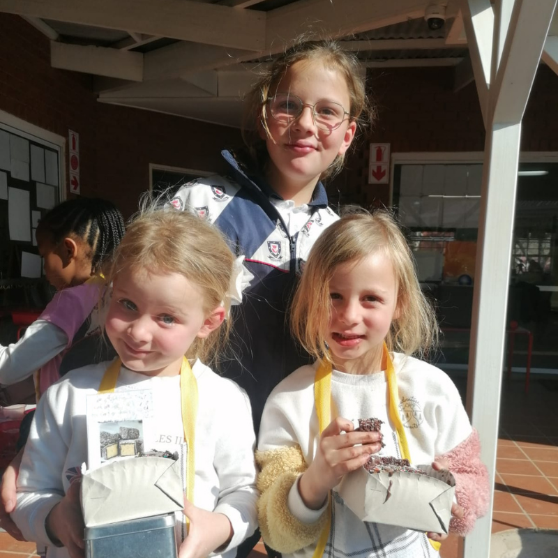 Three children standing outdoors, holding small paper crafts and smiling in bright sunlight.