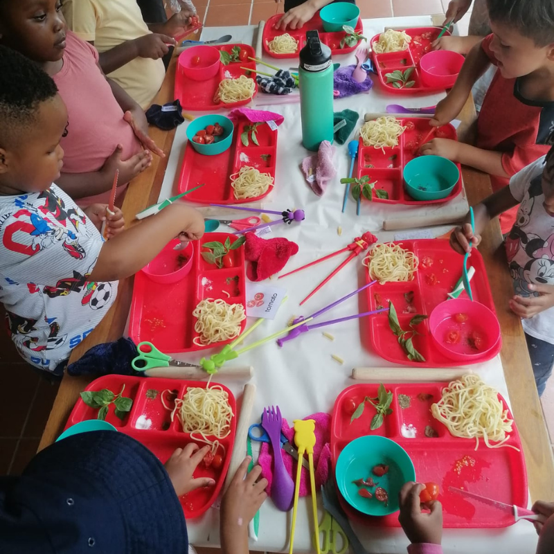 Children seated around a table eating spaghetti from red trays with pink and teal plates and cups