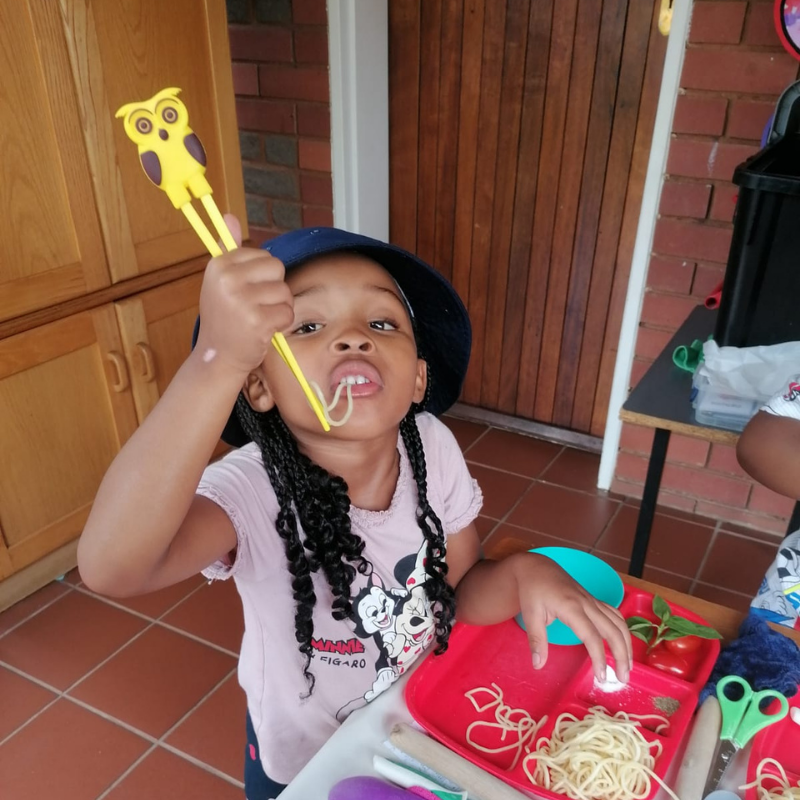 Child holding yellow tongs over a red tray of noodles indoors