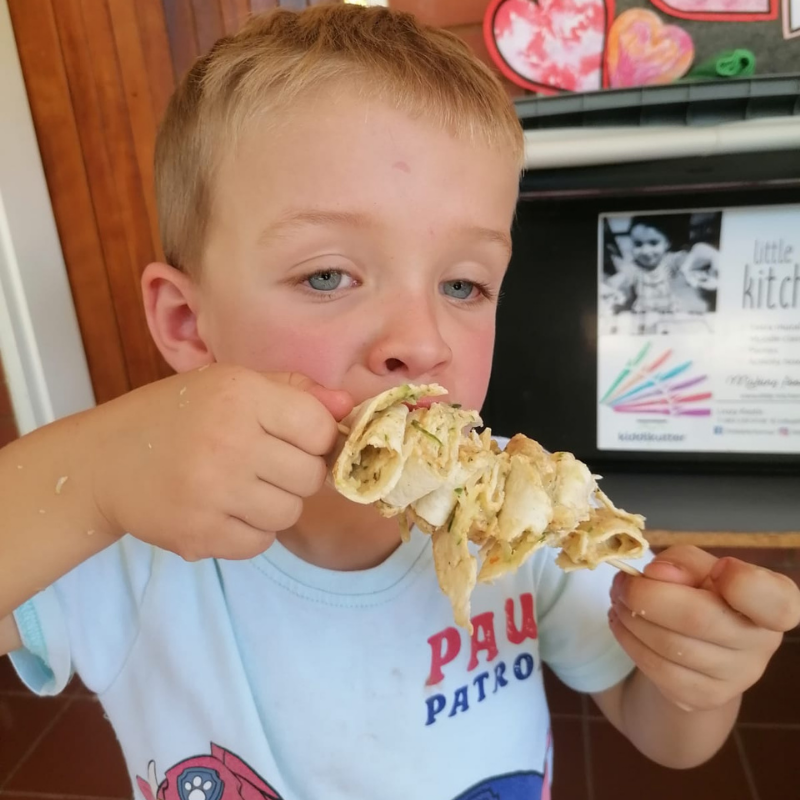 Child in a blue shirt eating a large folded sandwich at a kitchen table