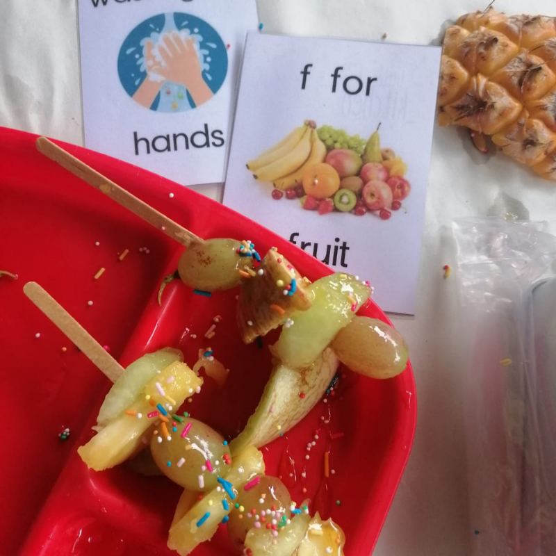 Fruit skewers with sprinkles on a red plate beside an “f for fruit” card and “hands” sign