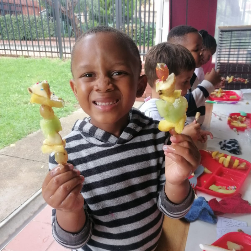 Smiling child holding two fruit skewers at a table, with other children and snacks in the background.