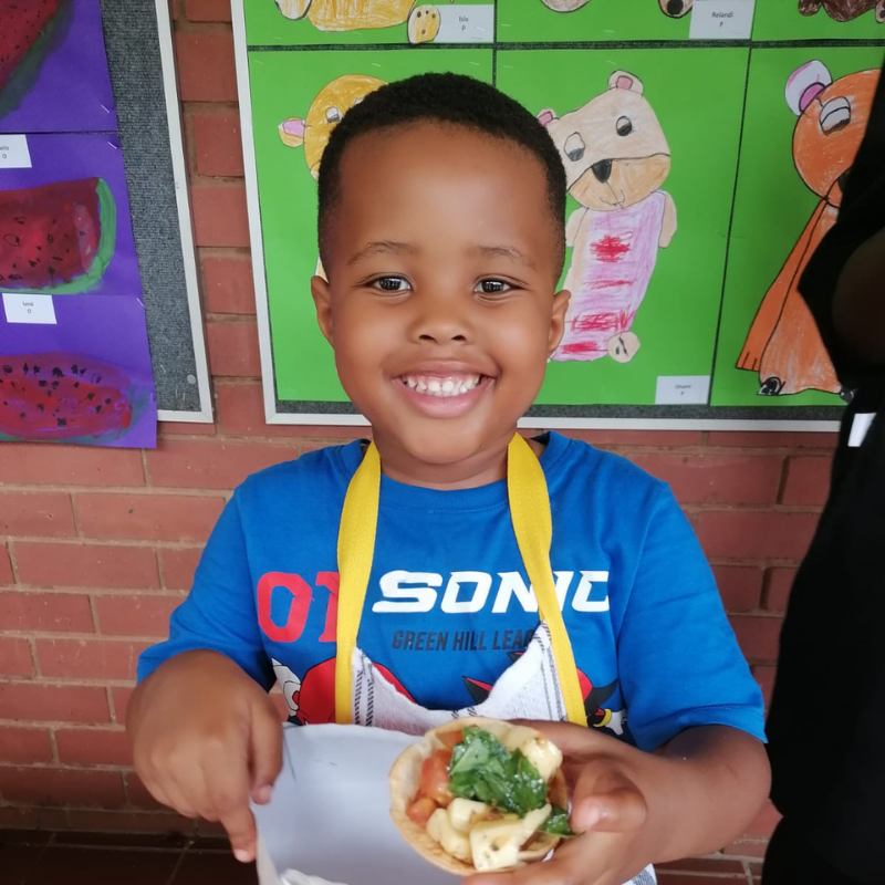 Smiling child holding a taco, wearing a blue Sonic shirt, standing in front of colorful classroom art.