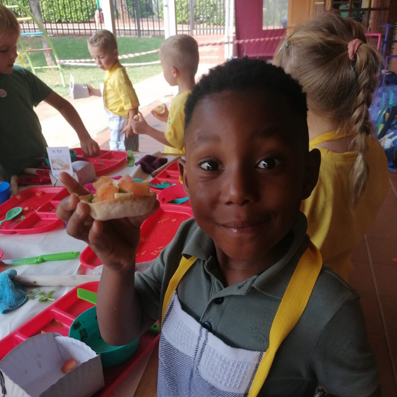 Child smiling and holding up a snack at a classroom table with art supplies and other children nearby