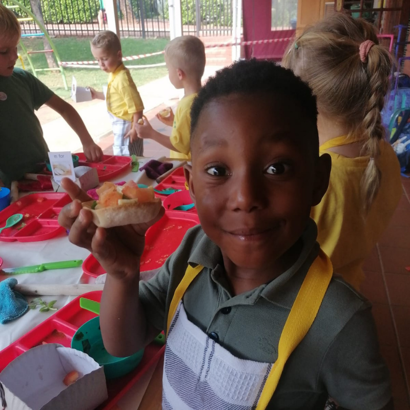 Child smiling and holding up a snack at a colorful cafeteria table with other children nearby
