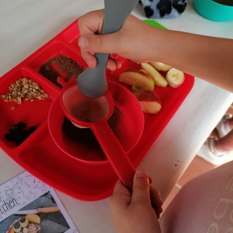 Hands using spoons to serve food from a red divided tray with small portions of fruit and snacks.