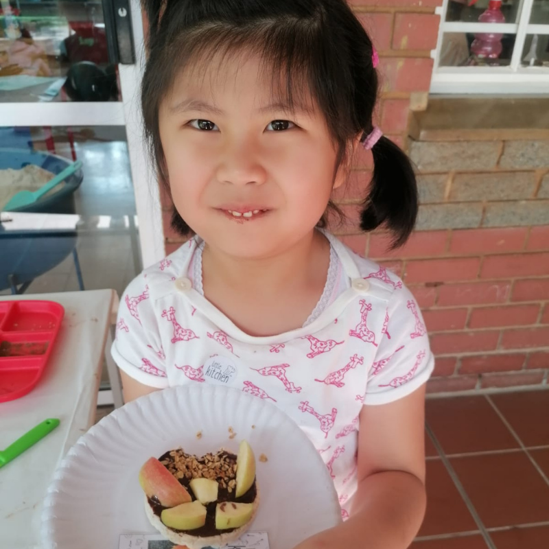 Young child holding a plate of sliced fruit and dessert, smiling at a table outdoors.