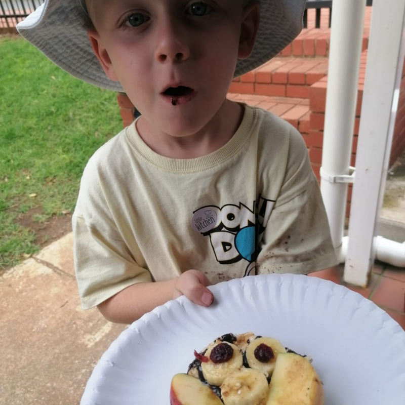 Child holding a plate of cookies outdoors, wearing a white hat and beige shirt with a surprised expression