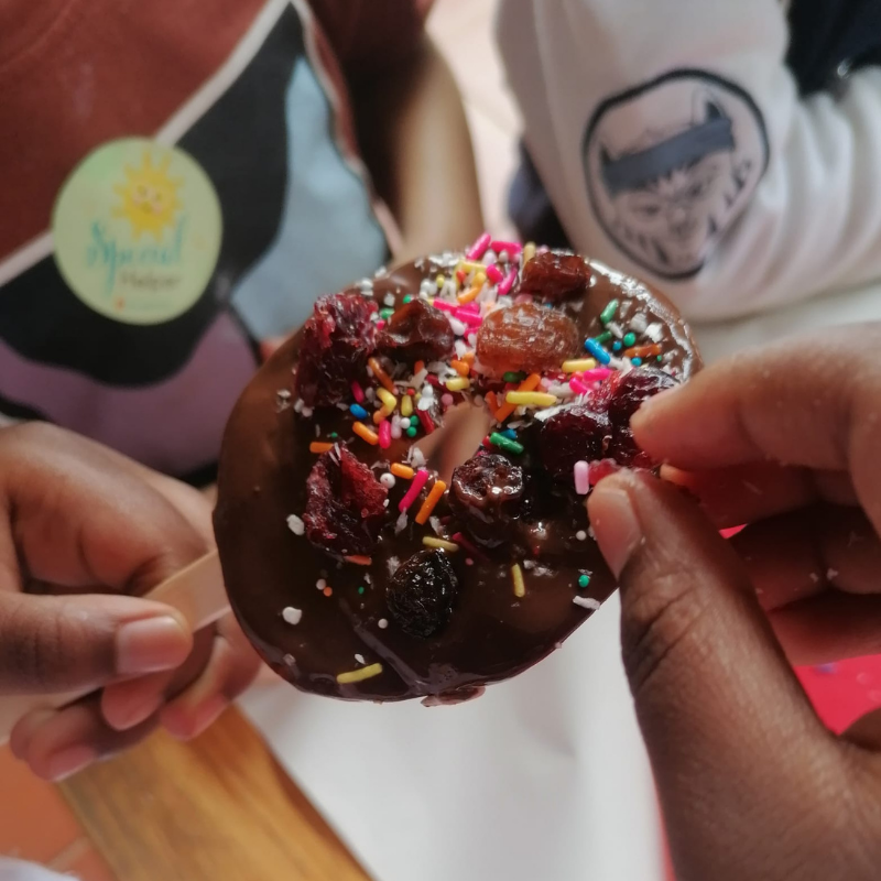 Hands holding a chocolate doughnut topped with colorful sprinkles and candy pieces