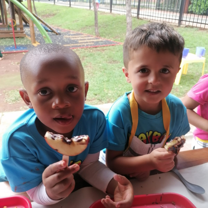 Two children at a picnic table eating snacks outdoors, with a playground in the background