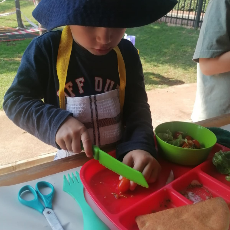 Child using a green knife to cut food at a red tray table outdoors, with scissors and a bowl nearby