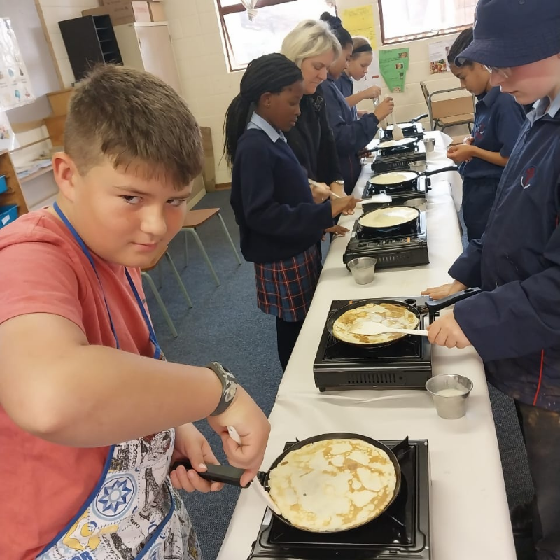 Students cooking pancakes on portable stoves in a classroom, with one boy stirring batter in the foreground