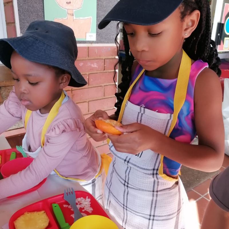 Two children in aprons and hats preparing food at a table outdoors