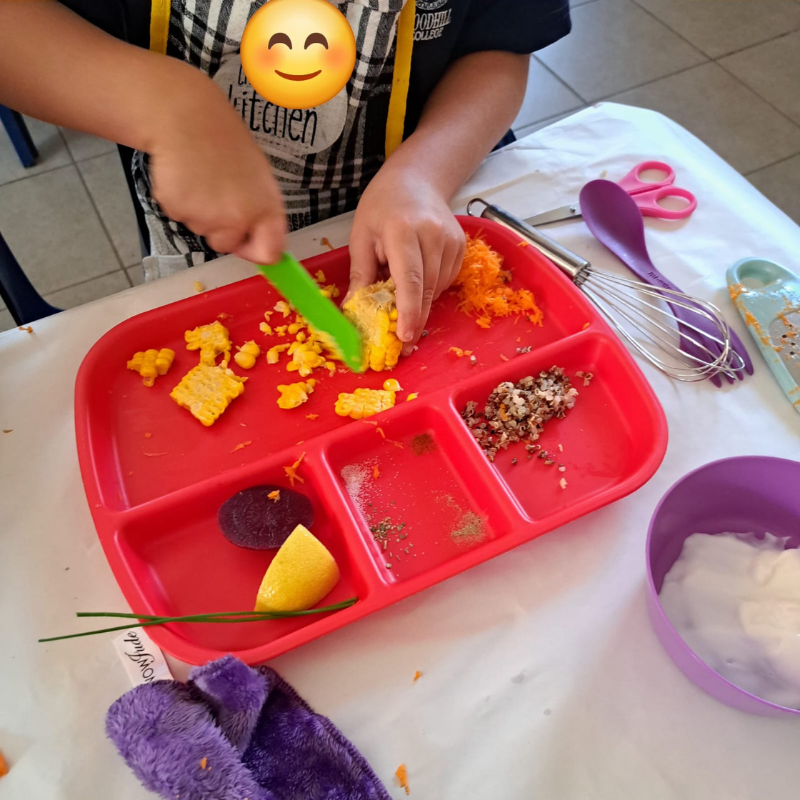 Child using a green spoon to mix food in a red divided tray on a table with utensils and cups
