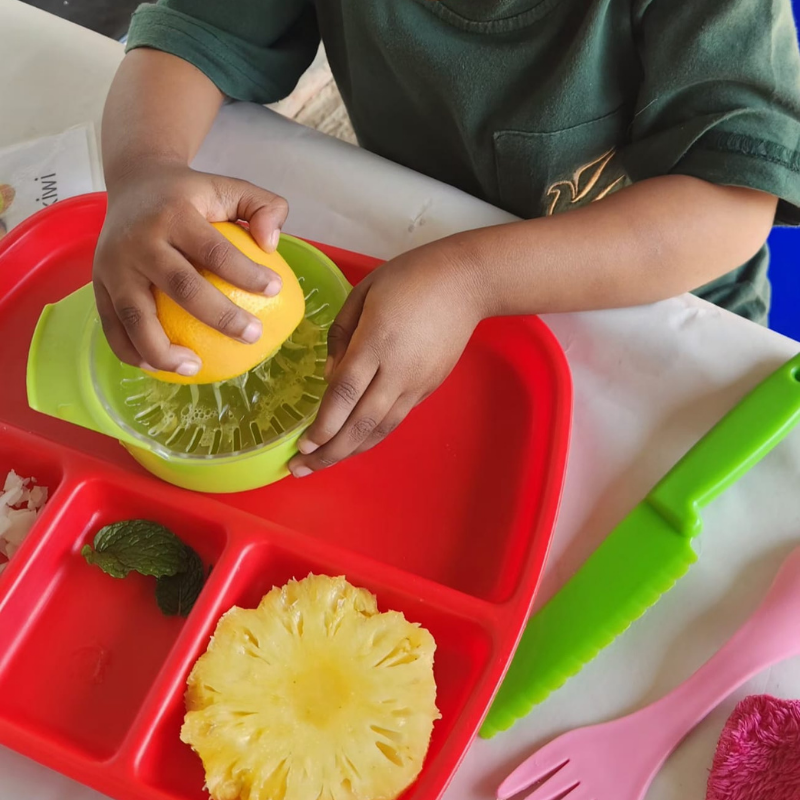 Child squeezing citrus into a green juicer on a red segmented tray with pineapple and lime pieces