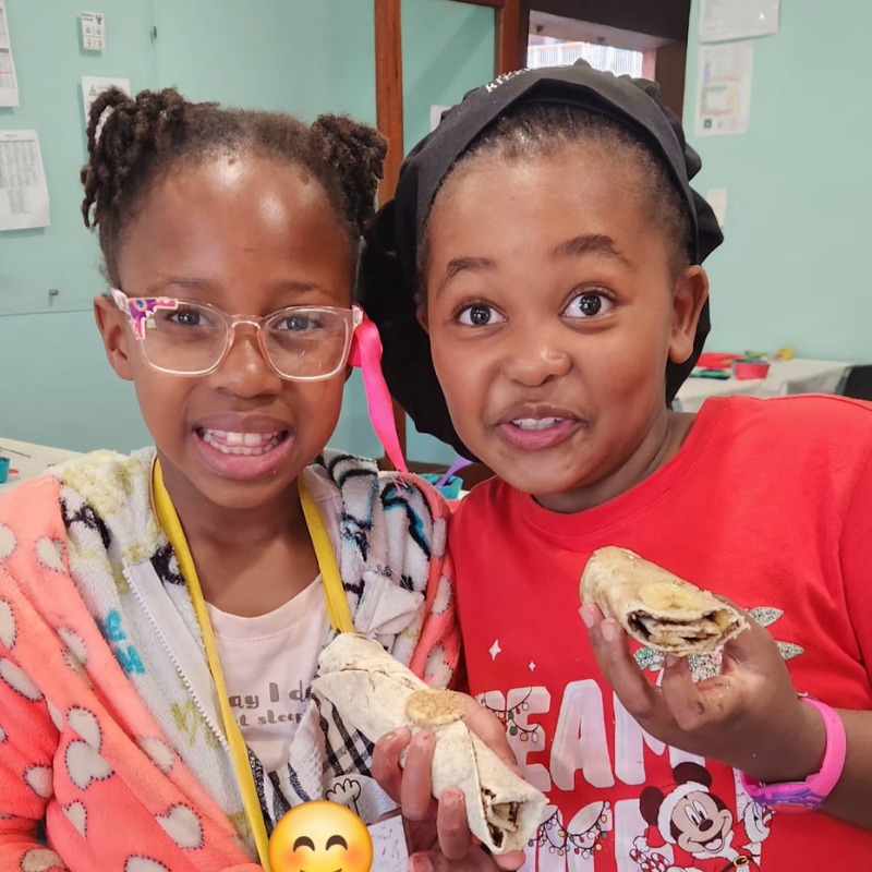 Two smiling children in a classroom holding small paper crafts, one in pink glasses and one in a red shirt.