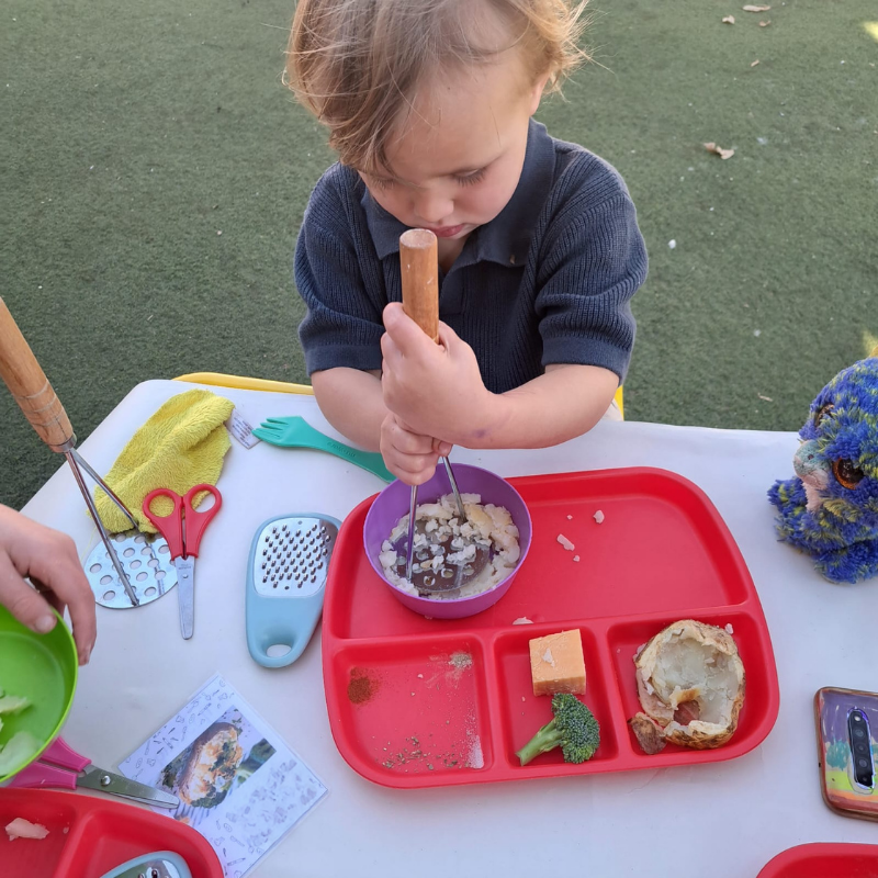Child using a wooden pestle to mash ingredients in a purple bowl on a red tray outdoors.