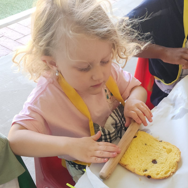 Child stirring dough with a rolling pin on a tray, wearing a pink shirt and yellow apron
