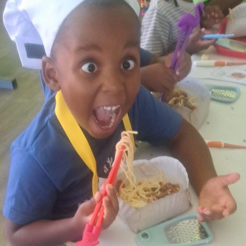 Child in blue shirt and white chef hat, laughing while holding red chopsticks at a table with noodles.