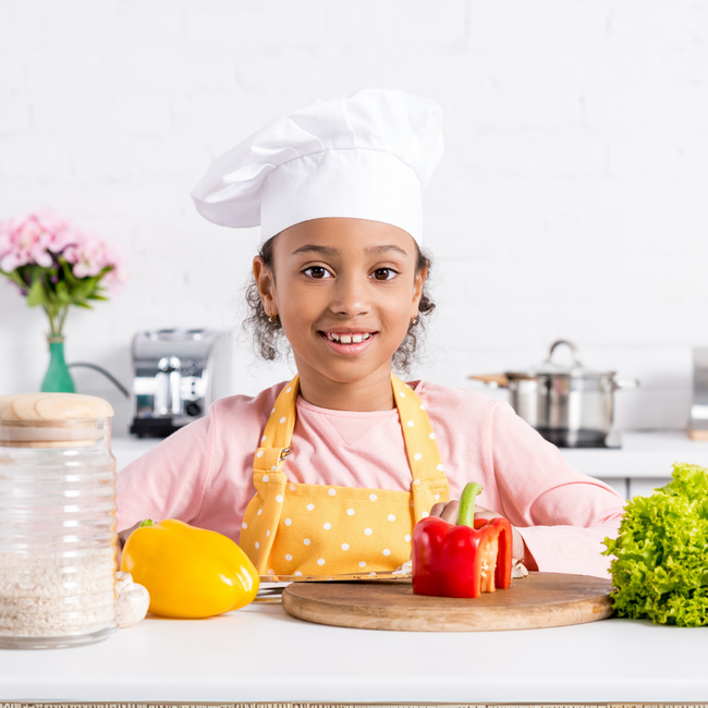 Child chef smiling in a kitchen, wearing a white hat and yellow apron beside fresh vegetables and a cutting board