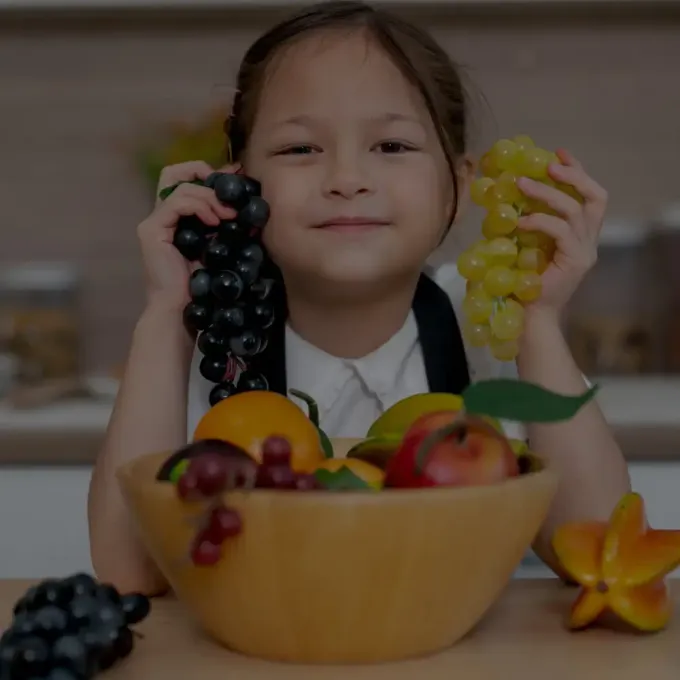 Child holding black and green grapes behind a bowl of mixed fruit on a table