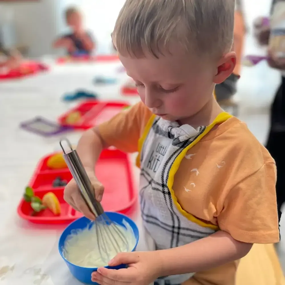 Child stirring a blue bowl with a whisk at a table, with a tray of fruit nearby.