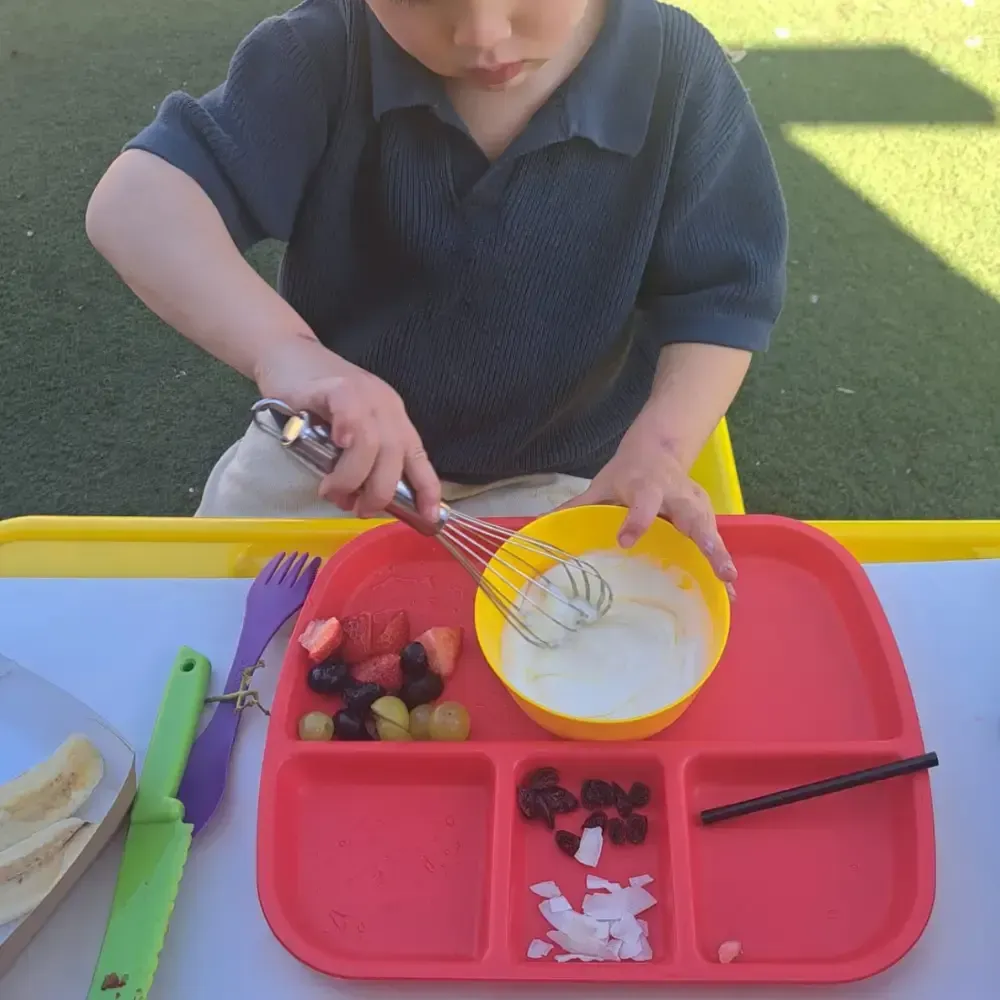 Child mixing batter in a yellow bowl at an outdoor play table with fruit and utensils.
