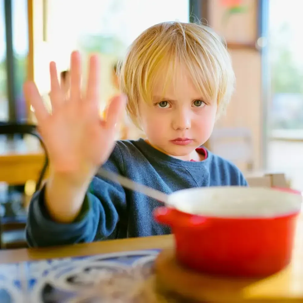 Child in a blue shirt raises a hand at a table with a red pot in a bright kitchen