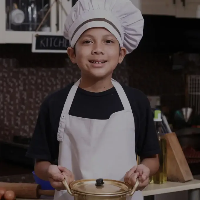 Smiling child in chef hat and apron holding a covered pot in a kitchen setting